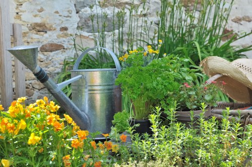 The image depicts a well-maintained garden corner featuring a vibrant flower bed of orange and yellow flowers in the foreground, complemented by lush green foliage and small shrubs behind. A weathered stone wall forms the backdrop, adding a rustic charm to the outdoor space. Central to the scene is a silver metal watering can with a curved handle and spout, placed alongside a traditional straw gardening hat resting on the ground. Surrounding these are various potted plants with leafy green foliage, some with delicate pink flowers, indicating a cultivated garden area suitable for professional landscaping and gardening services. The overall environment appears to be outdoors on a bright, possibly sunny day, with natural light illuminating the vibrant colours and textures of the plants and garden tools. The scene emphasizes a tidy, inviting garden environment that could be part of a residential outdoor space in Sudbury, aligning with Gardening Services Sudbury’s focus on lawn and garden maintenance, planting, and outdoor care expertise.