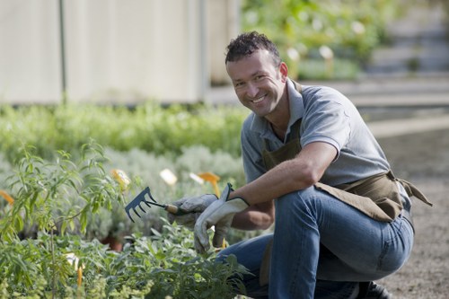 A smiling man kneels on a gravel pathway in a well-maintained garden, surrounded by lush green plants and flowering shrubs. He is wearing a grey shirt, blue jeans, and gardening gloves, and holds a small gardening fork while tending to the plant bed. Behind him, there is a wooden fence and a tiered garden with steps leading to a higher level, where additional greenery and foliage are visible. The garden features a mixture of dense grass, decorative plants, and freshly tilled soil, with natural daylight illuminating the scene. The setting appears to be a tidy and landscaped outdoor space, suitable for gardening and lawn care services in Sudbury, with a focus on healthy growth and ordered planting arrangements.
