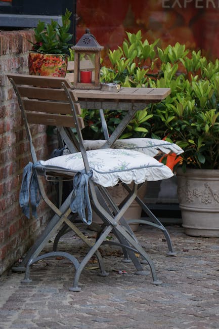 A small outdoor courtyard area featuring a weathered wooden bistro table and two matching chairs with tied fabric cushions, positioned against a red brick wall. The table is decorated with a decorativ
