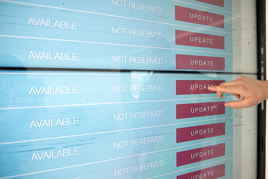 A person's hand pointing at a digital screen displaying an appointment schedule with the words 'AVAILABLE' and 'NOT RESERVED' in white text on a light blue background, alongside red 'UPDATE' buttons. 