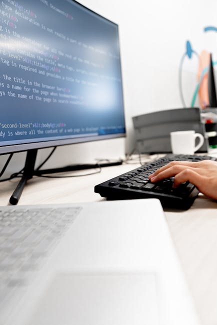 A close-up of a person's hand operating a black computer keyboard on a white desk, with a large monitor displaying lines of computer code in the background. The workspace includes a white coffee mug, 