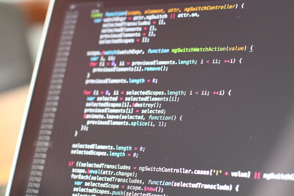 A close-up of a person's hand operating a black computer keyboard on a white desk, with a large monitor displaying lines of computer code in the background. The workspace includes a white coffee mug, 